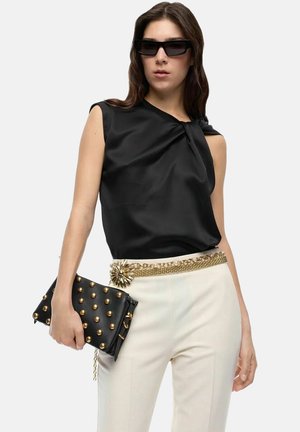 Donna con top nero senza maniche, pantaloni bianchi con cintura dorata decorativa, clutch nero borchiato e occhiali da sole rettangolari neri.