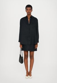 Black oversized button-up shirt paired with a black mini skirt. Model carries a textured black handbag and wears beige pointed-toe flats.