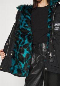 Svart parkas med en teal fuskpälsfoder som har ett leopardmönster, dragkedjestängning och silverfärgade detaljer.