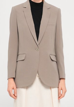 Beige getailleerde blazer met enkelvoudige knoopsluiting, klepzakken, gedragen over een zwarte top en witte broek.