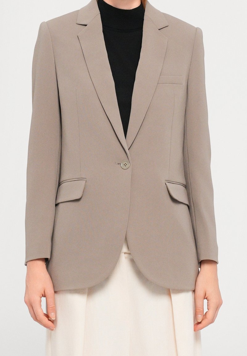 Blazer beige ajusté avec fermeture à un bouton, poches à rabat, porté sur un top noir et un pantalon blanc.