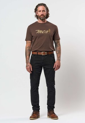 Moški z ramenami do dolgimi lasmi in tetovažami, ki nosi rjavo majico z grafiko, črne hlače, rjav pas in rjave škornje, stoji na belem ozadju.