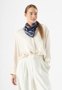 Écharpe en soie bleue avec des motifs complexes et des accents blancs, portée autour du cou, associée à un blouse crème et un pantalon blanc taille haute.