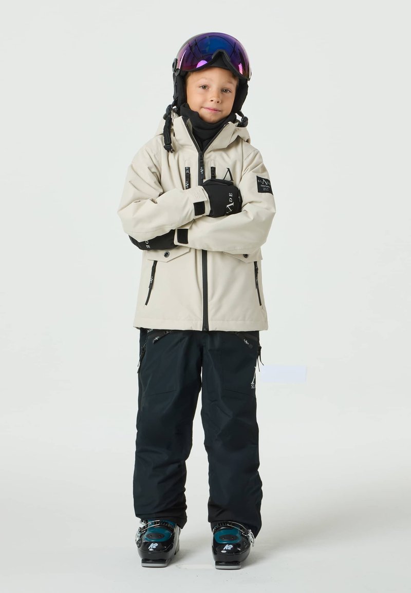 Ski outfit featuring a beige waterproof jacket with black accents, matching black pants, and winter boots with teal details.