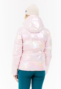 Puffjacka i iriserande ljusrosa material med huva, med sydda sektioner och en figurnära design. Bärs tillsammans med blågröna byxor.