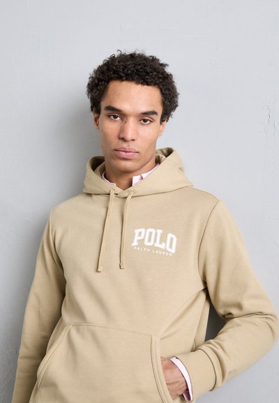 Haut de survêtement beige avec une poche frontale, arborant le texte blanc "POLO RALPH LAUREN". Matériau doux et cordon de serrage ajustable au niveau de la capuche.