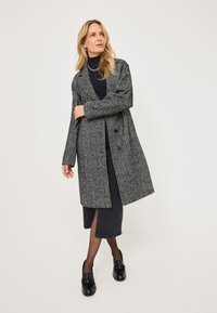 Houndstooth-patterned grey coat with a lapel collar, long sleeves, and two black buttons. Worn over a dark top and skirt. Black shoes.