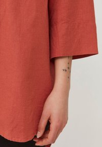 Rustfarbenes Leinenhemd mit Dreiviertelärmeln, das eine schlichte Textur präsentiert. Eine Hand ist sichtbar, die ein Tattoo am Handgelenk zeigt.