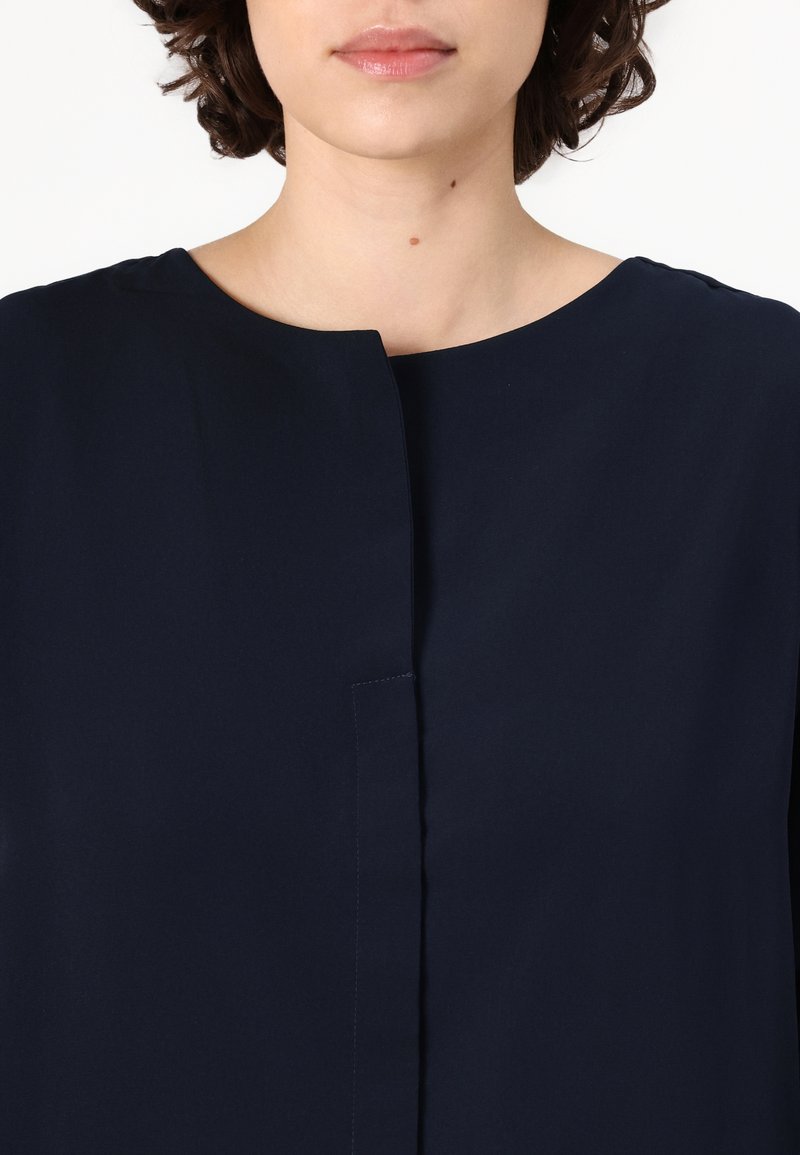 Marineblaue Bluse mit Rundhalsausschnitt und vorderem Knopfenverschluss. Der Stoff wirkt glatt mit einem dezenten Glanz und ohne sichtbare Muster oder Akzente.