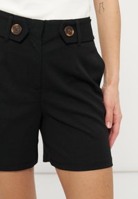 Short noir taille haute avec poches latérales avant et deux grands boutons marron sur la ceinture, porté avec un haut blanc, tatouage visible sur le bras.