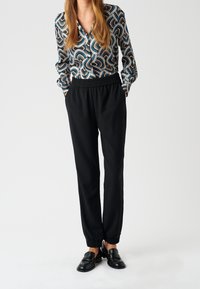 Pantalon noir à taille élastique avec jambes fuselées, associé à un chemisier boutonné à motifs ondulés multicolores. Des mocassins noirs complètent la tenue.