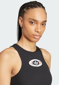Čierny rebrovaný tielko s bezrucným dizajnom a okrúhlym vyšitým logom v bielej a čiernej farbe. Má priliehavý strih a jemnú textúru.