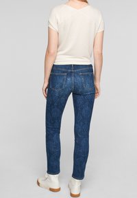Jeans de mezclilla de color azul oscuro con un intrincado patrón de paisley, combinados con una blusa de manga corta de canalé color crema y botas blancas hasta el tobillo.