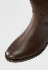 Brązowe skórzane botki za kostkę o gładkiej fakturze, zaokrąglonym nosie i subtelnym haftowaniu wzdłuż górnej szwy. Solidna czarna podeszwa.