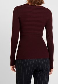 Haut bordeaux à manches longues avec texture rayée et ourlet côtelé. Caractérisé par une silhouette ajustée et une finition lisse, associé à un pantalon à fines rayures.