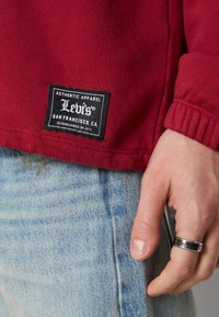Camicia rossa a maniche lunghe in cotone caratterizzata da un'etichetta nera Levi's con testo bianco, abbinata a jeans in denim blu sbiaditi. La mano indossa un anello in argento.
