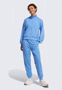 Svetlo modra atletska trenerka iz gladkega materiala, ki vsebuje jakno z zadrgo in ovratnikom ter ujemajoče se trenirke, okrašene z logotipom.