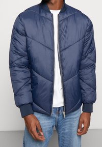 Navyblauwe pufferjas met een rits aan de voorkant, ribbels aan de mouwen en een chevron stikpatroon; heeft een lichte, glanzende stofafwerking.