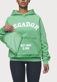 Sudadera verde con texto blanco "EGADOR" y "NEXT WAVE IS NOW", con un ajuste relajado y puños acanalados, que muestra una textura suave.