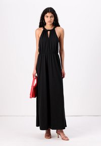 Vestuário maxi preto com decote halter, cintura franzida, detalhe em forma de olho na frente, combinado com uma bolsa vermelha e sandálias de tiras.