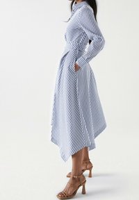 Vestido camisa largo a rayas azules y blancas con mangas largas, cuello, bolsillos laterales y un dobladillo asimétrico. Combinado con sandalias marrones de tacón.