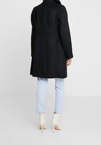 Černý vlněný kabát s ušitým designem, střední délky, s vysokým límečkem a knoflíkovými manžetami. Nesený přes světle modré kalhoty a béžové lodičky.