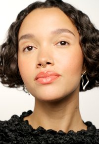 Portrait d'une personne avec des cheveux courts et bouclés, portant un haut noir à volants, un anneau septum en or et des boucles d'oreilles ondulées en argent. Peau lisse, lèvres rose pâle.