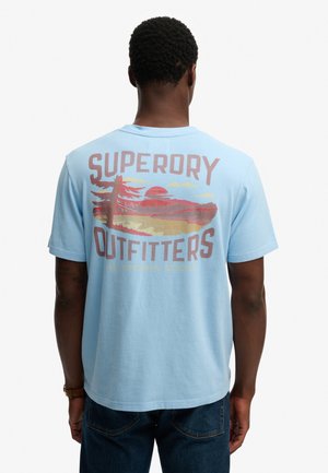 Hombre con camiseta azul clara con el gráfico del atardecer y árbol de "Superdry Outfitters" en la espalda, combinado con vaqueros azul oscuro.