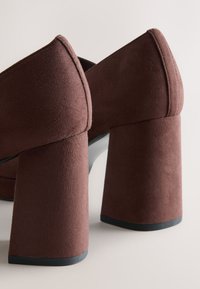 Bruna mockaskor med hög klack, fyrkantig tå och kraftig klack. Ytan är texturerad och klacken har en svart bas för stabilitet.