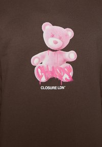 T-shirt marrone con una grafica di un orsetto rosa con gocce e il testo “CHAOS” in stile graffiti, sotto il nome del brand “CLOSURE LDN.”