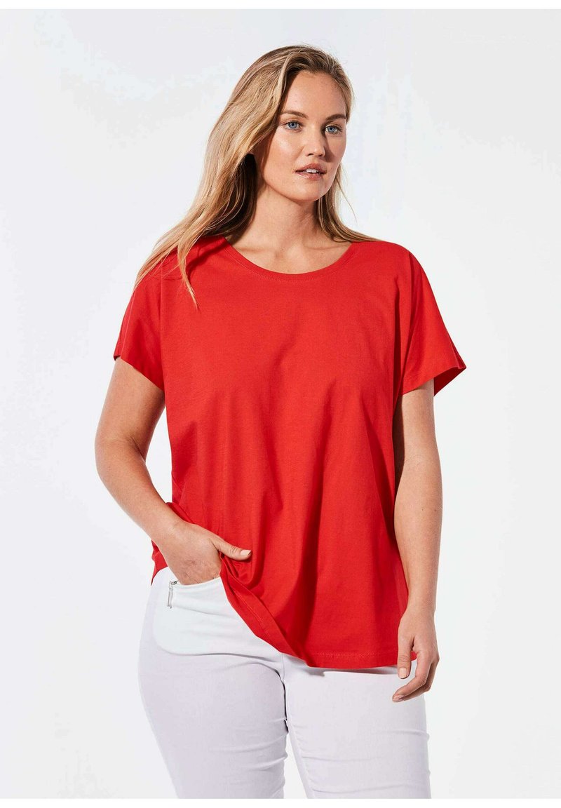 Rotes Baumwoll-T-Shirt mit kurzen, weiten Ärmeln und rundem Ausschnitt. Mit einem gebundenen Detail an der Seite, kombiniert mit weißen Hosen.