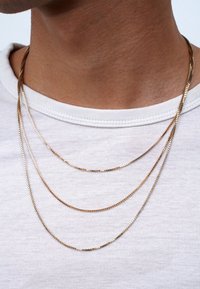 Goldfarbene, dreilagige Kettenhalskette mit eleganten, flachen Gliedern. Über einem weißen Rundhalsausschnitt-Shirt getragen, was ihren Glanz und ihre Textur betont.