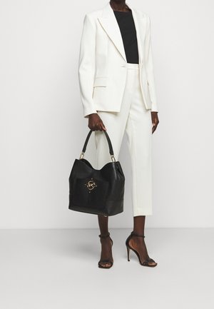 Zwarte leren handtas met een gestructureerde afwerking, voorzien van een gouden logo-accent en een gebogen vorm. Model in een witte suit met een zwarte top.