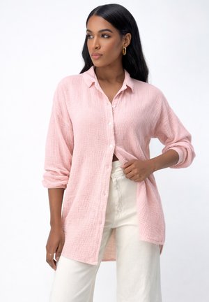 Frau mit langen dunklen Haaren, die ein hellrosa strukturiertes Hemd mit Knöpfen und cremeweiße Hose trägt, schaut mit neutralem Gesichtsausdruck nach links.