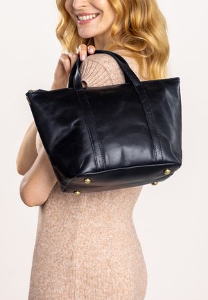 Black leather tote bag with two sturdy handles, rectangular shape, embossed logo, and brass feet on the bottom for stability.