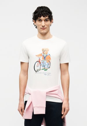 Mladý muž s kučeravými vlasmi, oblečený v bielom tričku s potlačou medveďa na bicykli, so ružovou mikinou previazanou okolo pása.