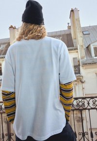 Bijela majica složena preko žute i smeđe prugaste majice dugih rukava. Model nosi crnu kapu.