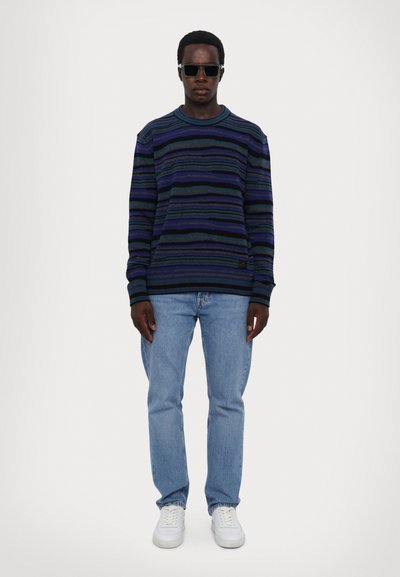 Uomo che indossa occhiali da sole neri, una maglia a strisce blu e nere, jeans azzurri e sneakers bianche, in piedi davanti a uno sfondo bianco semplice.