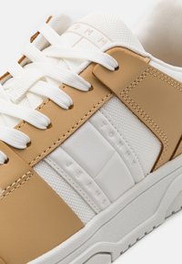 Baskets blanches et beige avec lacets blancs, panneaux en tissu texturé, empiècements en cuir et semelle en caoutchouc portant le nom de la marque "Tommy Jeans".