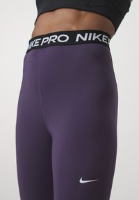Leggings atléticos de talle alto morados con una banda elástica negra que presenta "NIKE PRO" en texto blanco y un pequeño logo de la swoosh de Nike en blanco.