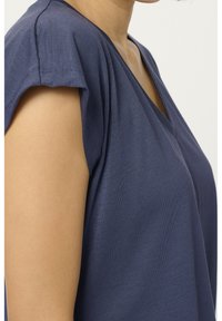 Persona che indossa una maglietta a maniche corte, di colore blu navy, con scollo a V, che mostra la spalla e la parte superiore del torso su uno sfondo semplice.