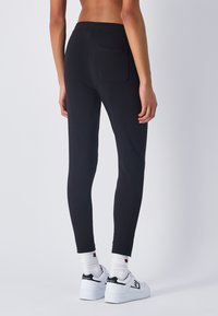 Pantalones de chándal negros con cintura elástica, ajustados y con un bolsillo trasero. Combinados con zapatillas blancas y calcetines con un logo.