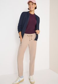 Marinblå zip-up jacka över en vinröd ribbad topp, beige byxor med snörning och vita sneakers med ljusblå accenter. Rosa keps på huvudet.