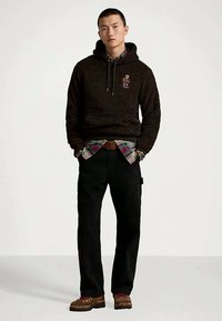 Hombre de pie con las manos en los bolsillos, vistiendo un sudadera de fleece negra con un logo de oso, camisa de cuadros, pantalones negros y zapatos marrones con cordones rojos.