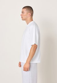 Bela majica s kratkimi rokavi, ohlapnega kroja z okroglim izrezom, v kombinaciji z sivimi trenirkami. Gladek teksturni material, minimalističen dizajn.