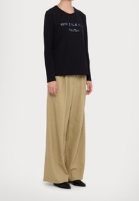 Persona che indossa una maglietta nera a maniche lunghe con testo applicato di paillettes e pantaloni larghi beige, in piedi di fronte a uno sfondo bianco.