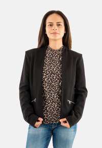 Femme avec de longs cheveux bruns portant un blazer noir, un haut à imprimé léopard et un jean bleu devant un fond blanc uni.