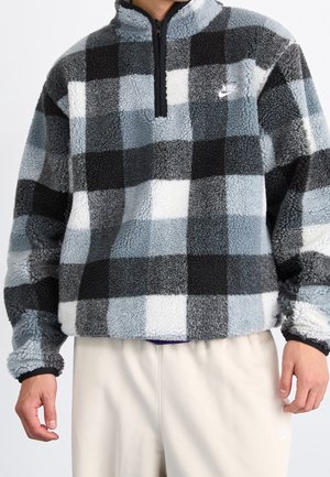 Personne portant un pull polaire à carreaux noir, gris et blanc avec une fermeture éclair d'un quart et le logo Nike sur la poitrine, associé à un pantalon de couleur claire.