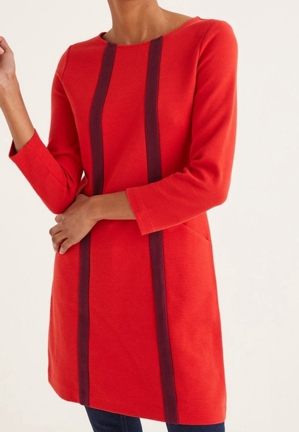 Femme portant une robe tunique rouge à manches longues avec deux bandes verticales foncées sur le devant, associée à un pantalon foncé.