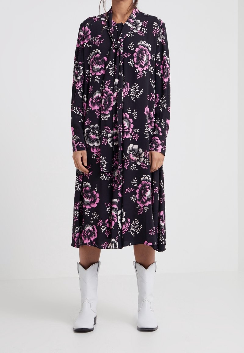 Femme portant une robe midi noire avec des motifs floraux roses et blancs et des bottes de cowboy blanches, debout devant un fond uni.
